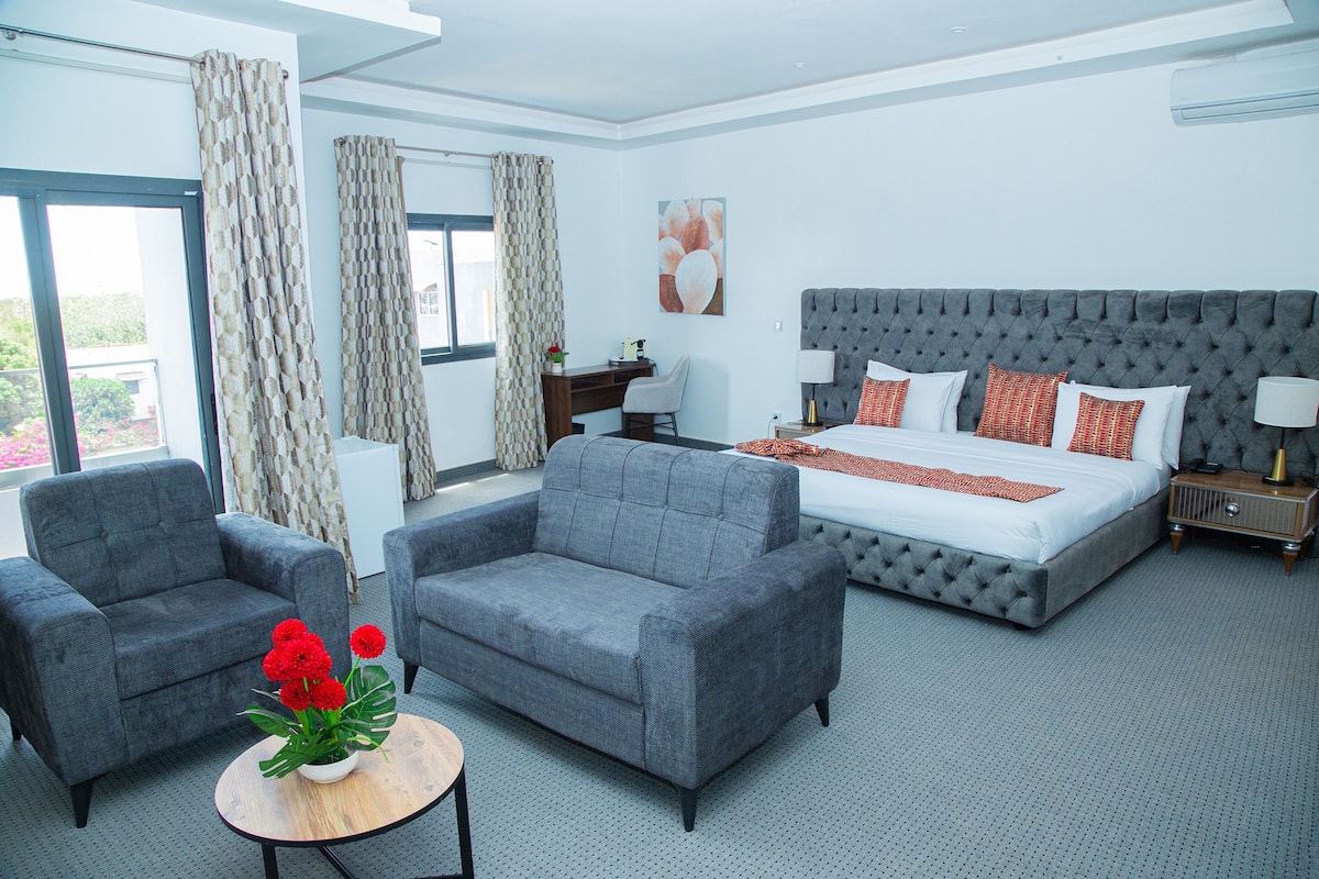 Une chambre d’hôtel moderne avec un grand lit, deux fauteuils, une petite table agrémentée de fleurs et une vue sur la verdure par les fenêtres.