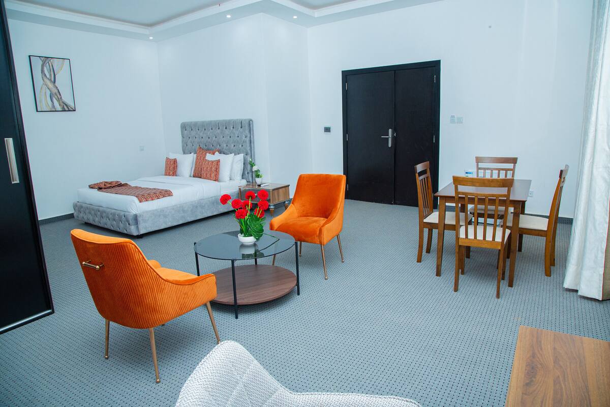 Une chambre d’hôtel avec un lit, deux fauteuils orange, une table ronde et un ensemble de mobilier pour la salle à manger.