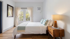 Un dormitorio con cama, mesita de noche y lámpara. Se observa una ventana con cortinas, un equipo de aire acondicionado y un ventilador de techo.