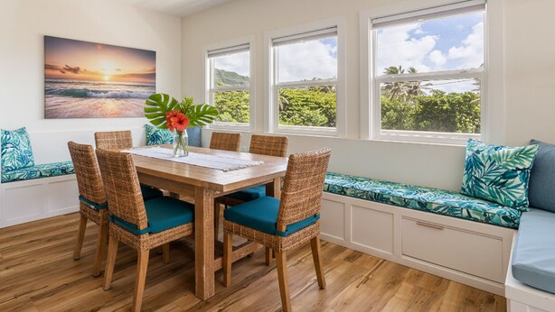 Un comedor con una mesa de madera y sillas de mimbre, vistas al exterior a través de amplios ventanales, y un banco con cojines.