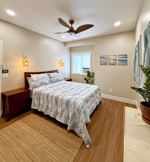 Un dormitorio con cama, mesitas de noche, ventilador de techo, luces empotradas en la pared, una ventana con cortinas, una planta en maceta y una obra de arte en la pared.