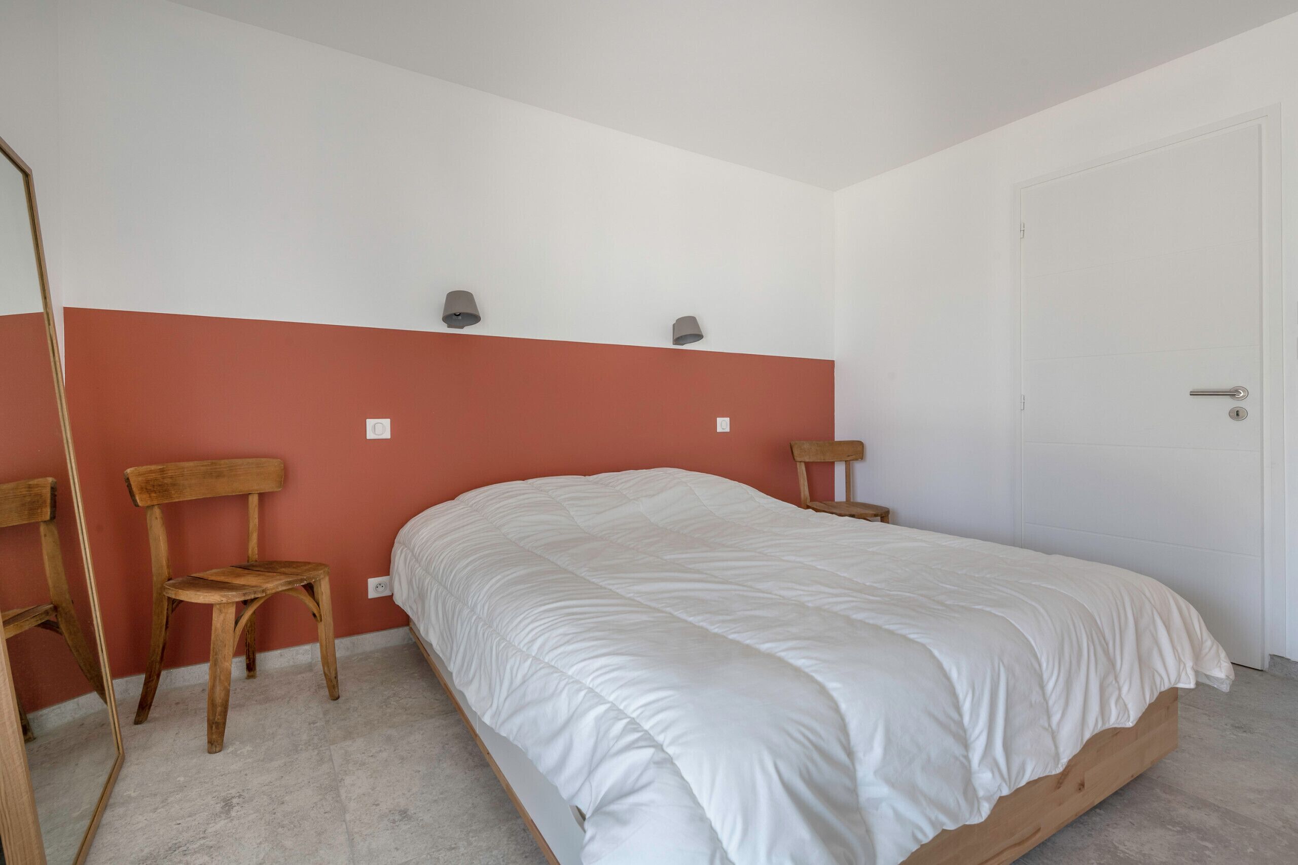 Een slaapkamer met een bed, twee stoelen, een spiegel en een witte deur.