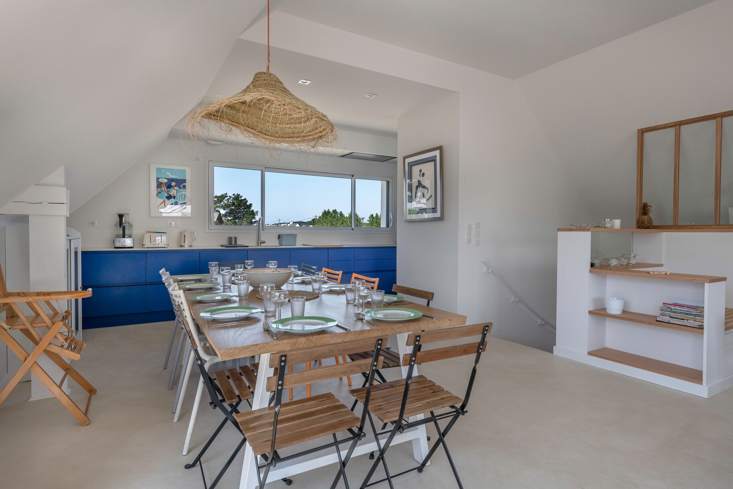 Une cuisine moderne avec des meubles bleus, un coin repas avec des tables et des chaises en bois, et une grande fenêtre donnant sur l’extérieur.