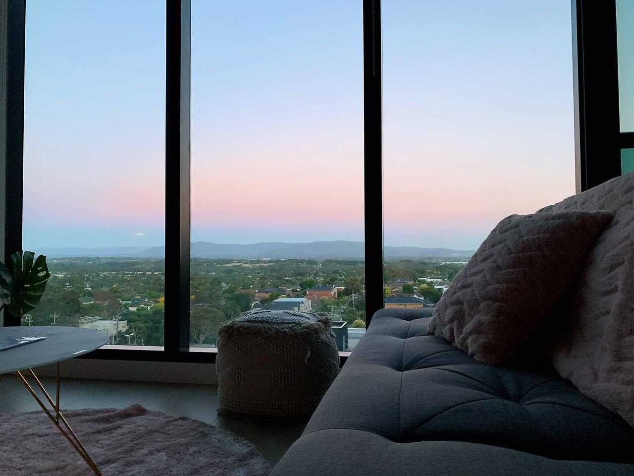 A modern living room with a large window, a grey sofa, a small ottoman, and a table with a plant.