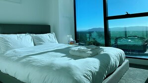A hotel room with a large bed, a nightstand, and a window offering a view of a landscape. - Skygarden Luxury 2-Bed, 2-Bath Apartment with Free Parking in Central Location (Glen Waverley)