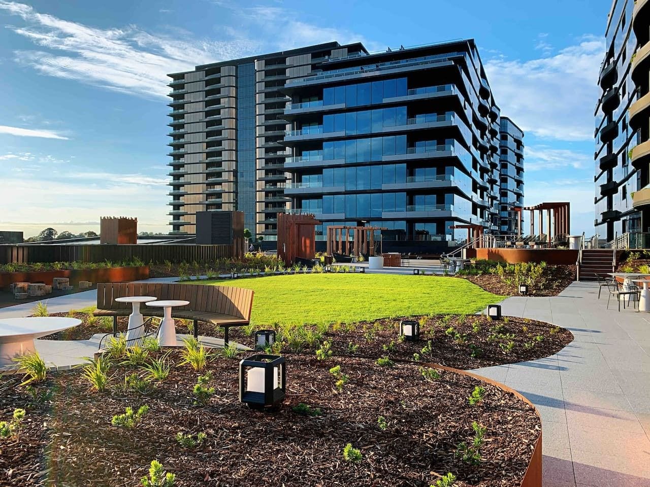 A modern building with multiple balconies, a central courtyard with grass and seating, and a clear sky.