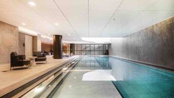 A modern indoor swimming pool with a clear glass barrier, a lounge area with seating, and a high ceiling with recessed lighting.