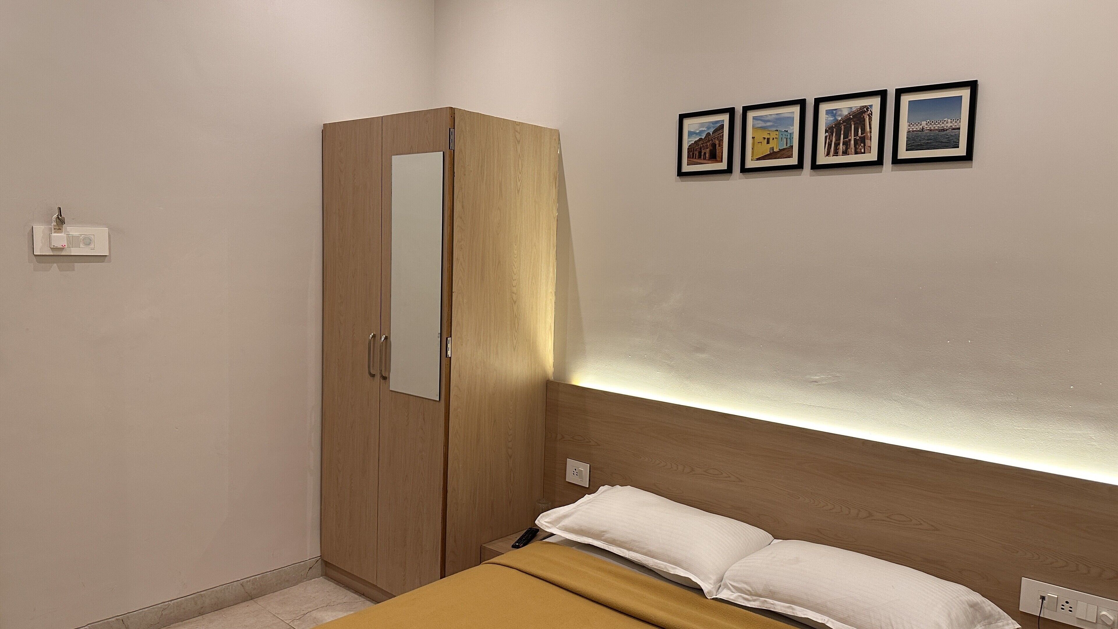 Une chambre avec une armoire en bois, un lit avec une literie blanche et une lampe fixée au mur au-dessus du lit. Quatre tableaux encadrés ornent le mur.