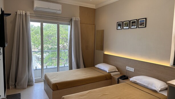 Une chambre d’hôtel avec deux lits, une grande fenêtre avec des rideaux, un appareil de climatisation, une table de chevet avec un téléphone et des tableaux encadrés au mur.