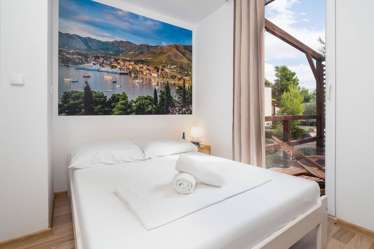 A hotel room with a bed, a nightstand, a window with curtains, and a painting of a coastal town.