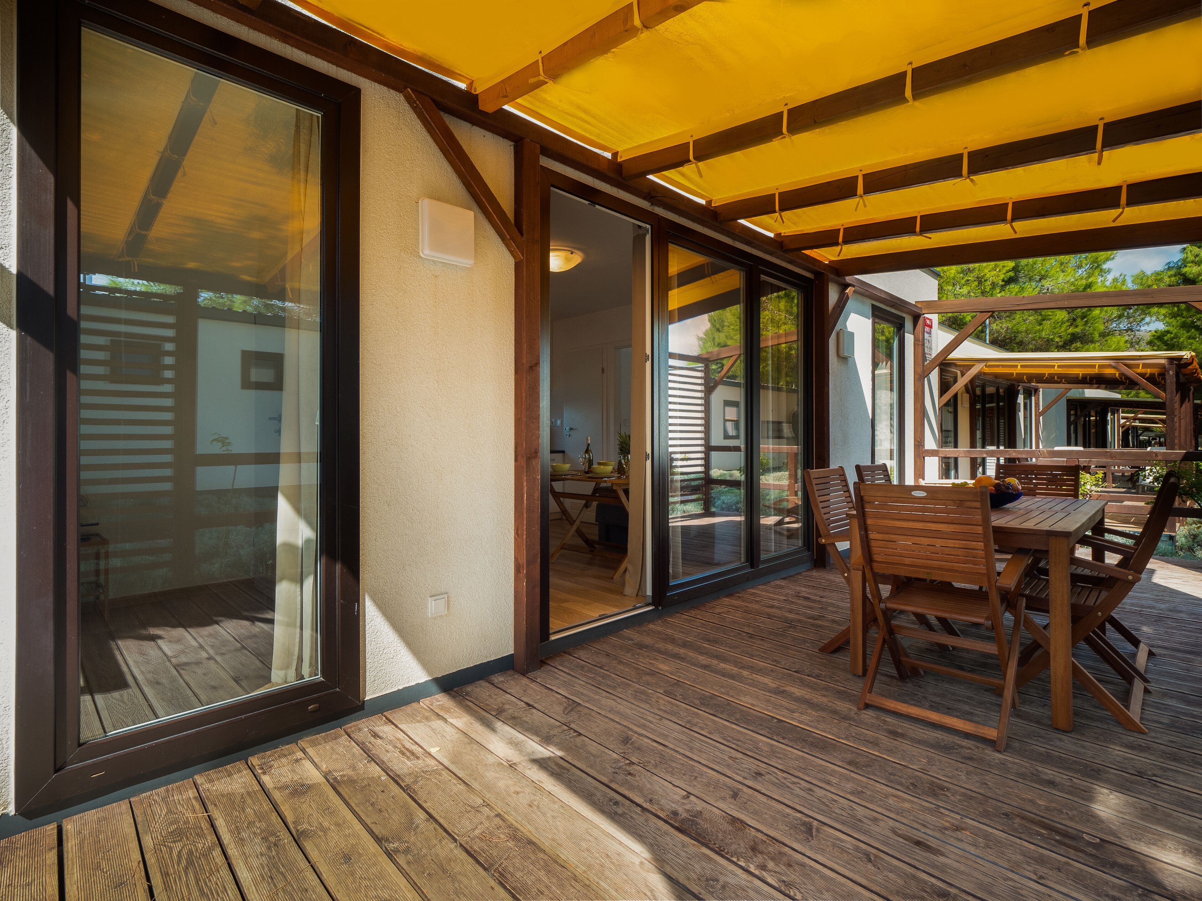 Un patio techado con suelo de madera, una mesa con sillas y un ventanal de vidrio que da a un espacio exterior.