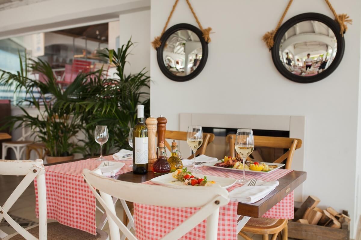 A dining table set with a checkered tablecloth, wine glasses, and bottles, surrounded by white chairs.