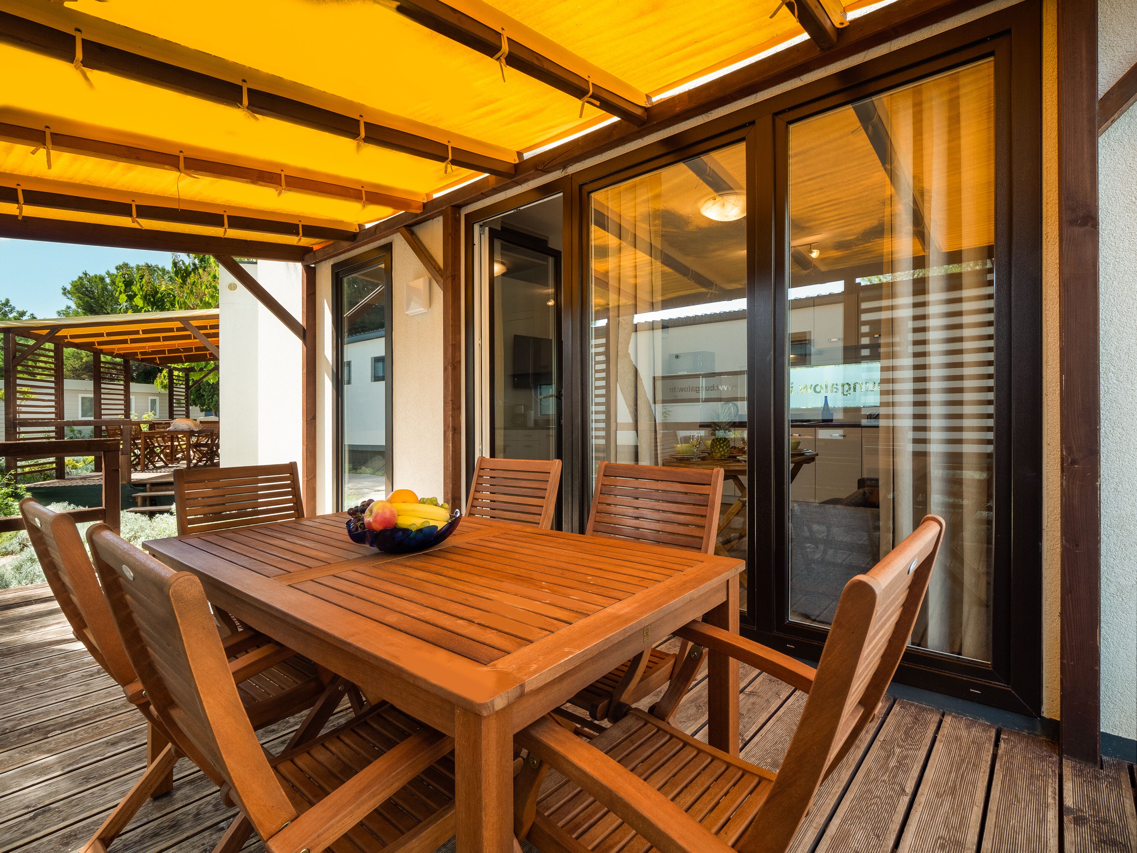An outdoor wooden deck with a table and chairs, a bowl of fruit, and a glass door leading to a kitchen area.