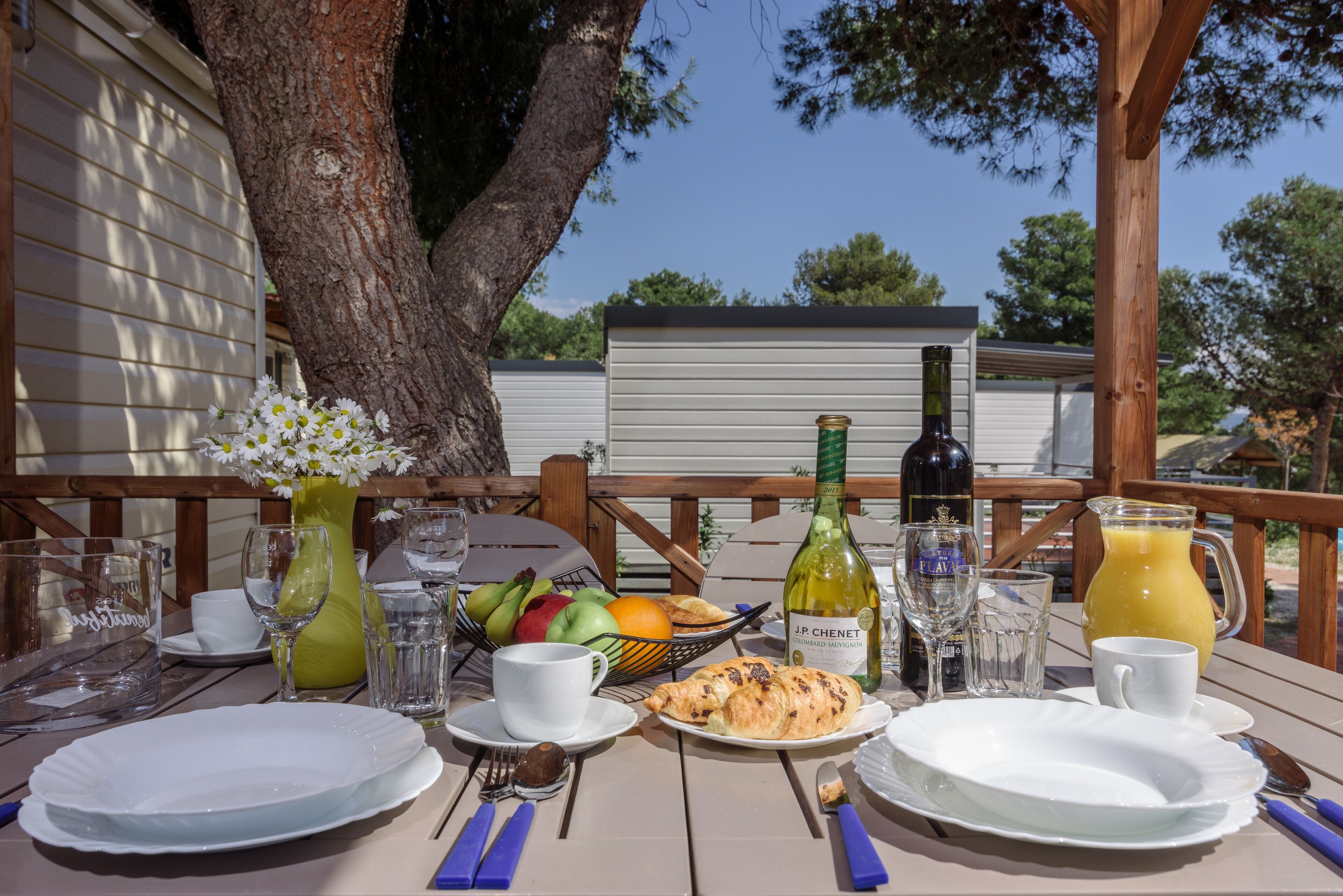 An outdoor dining table set with plates, cutlery, a bottle of wine, a pitcher of orange juice, and a basket of fruits.