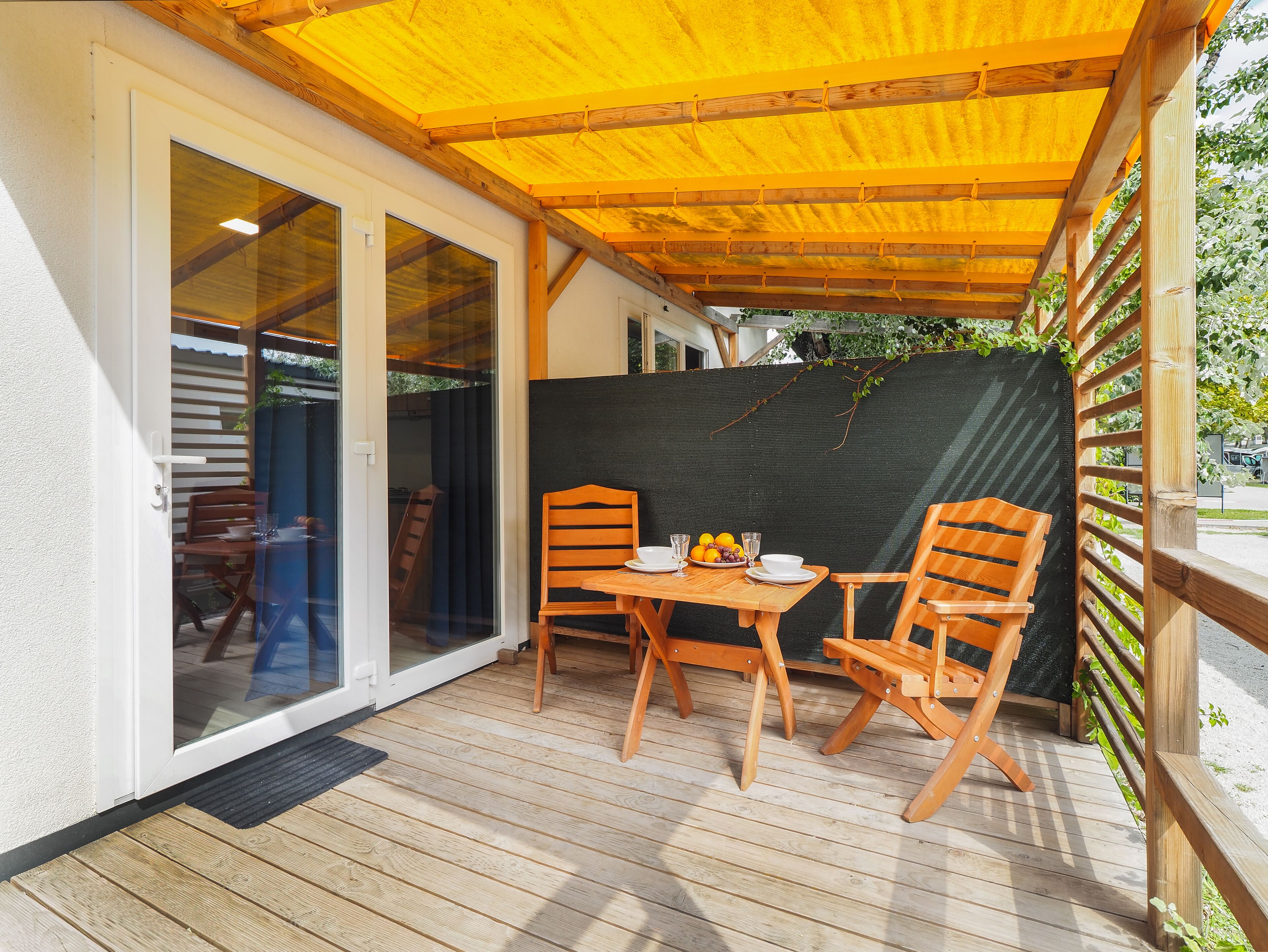Eine überdachte Terrasse mit Tisch und Stühlen, Holzboden und einer Glastür, die zu einem Innenbereich führt.
