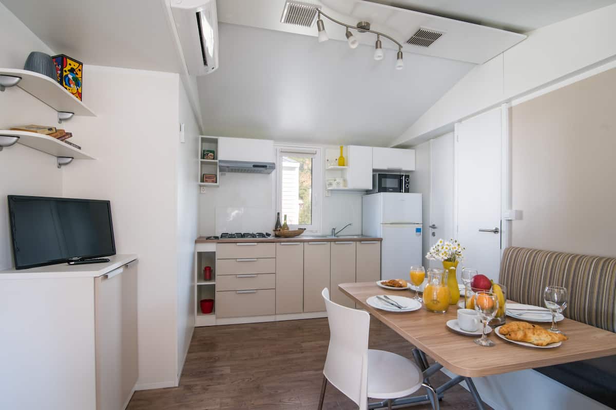 A modern kitchen with a dining area, white cabinetry, a flat-screen TV, and a breakfast setup with croissants and fruit.