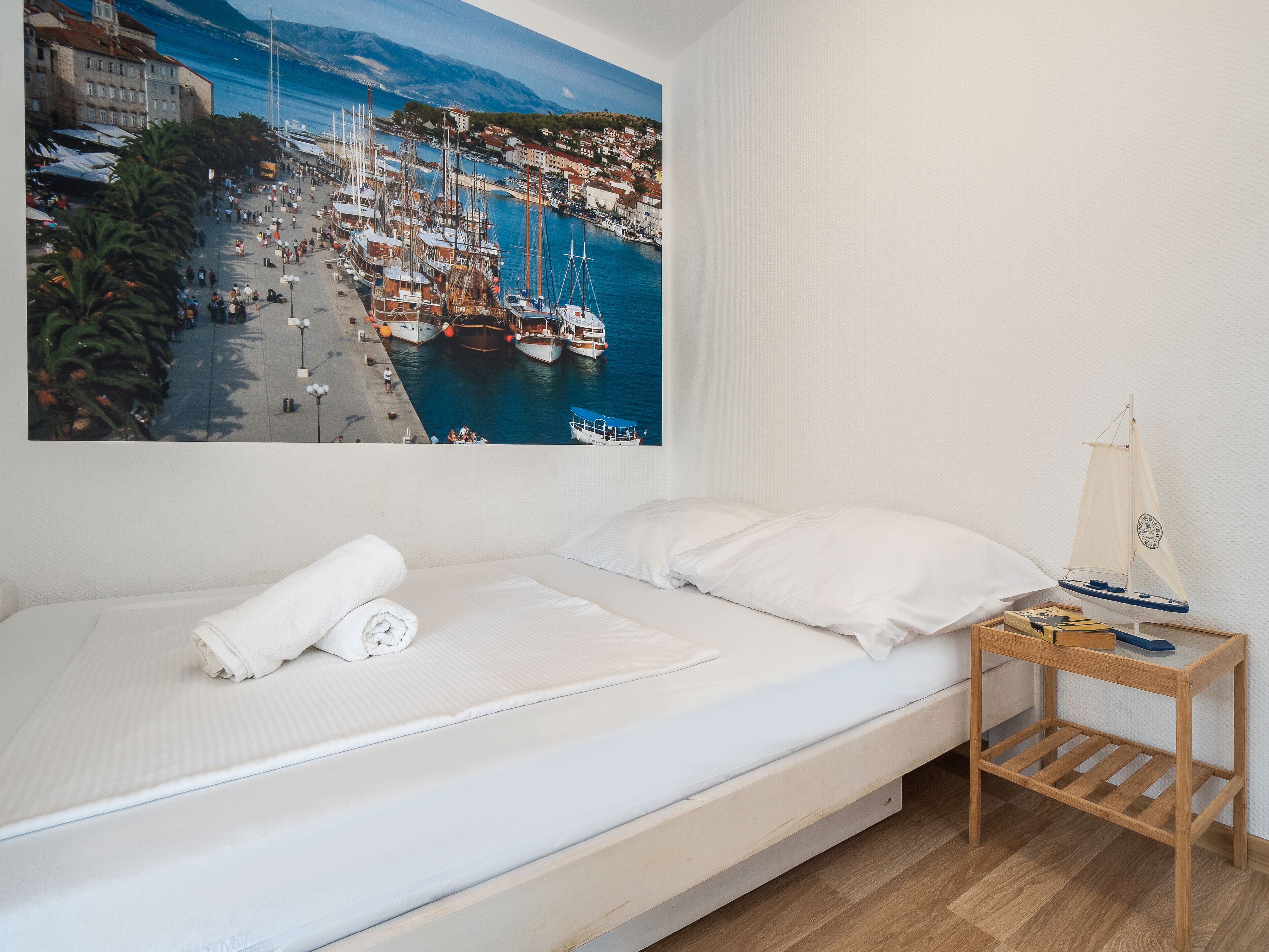 Een bed met witte lakens, een nachtkastje van hout met een model zeilboot, en een schilderij van een jachthaven aan de muur.