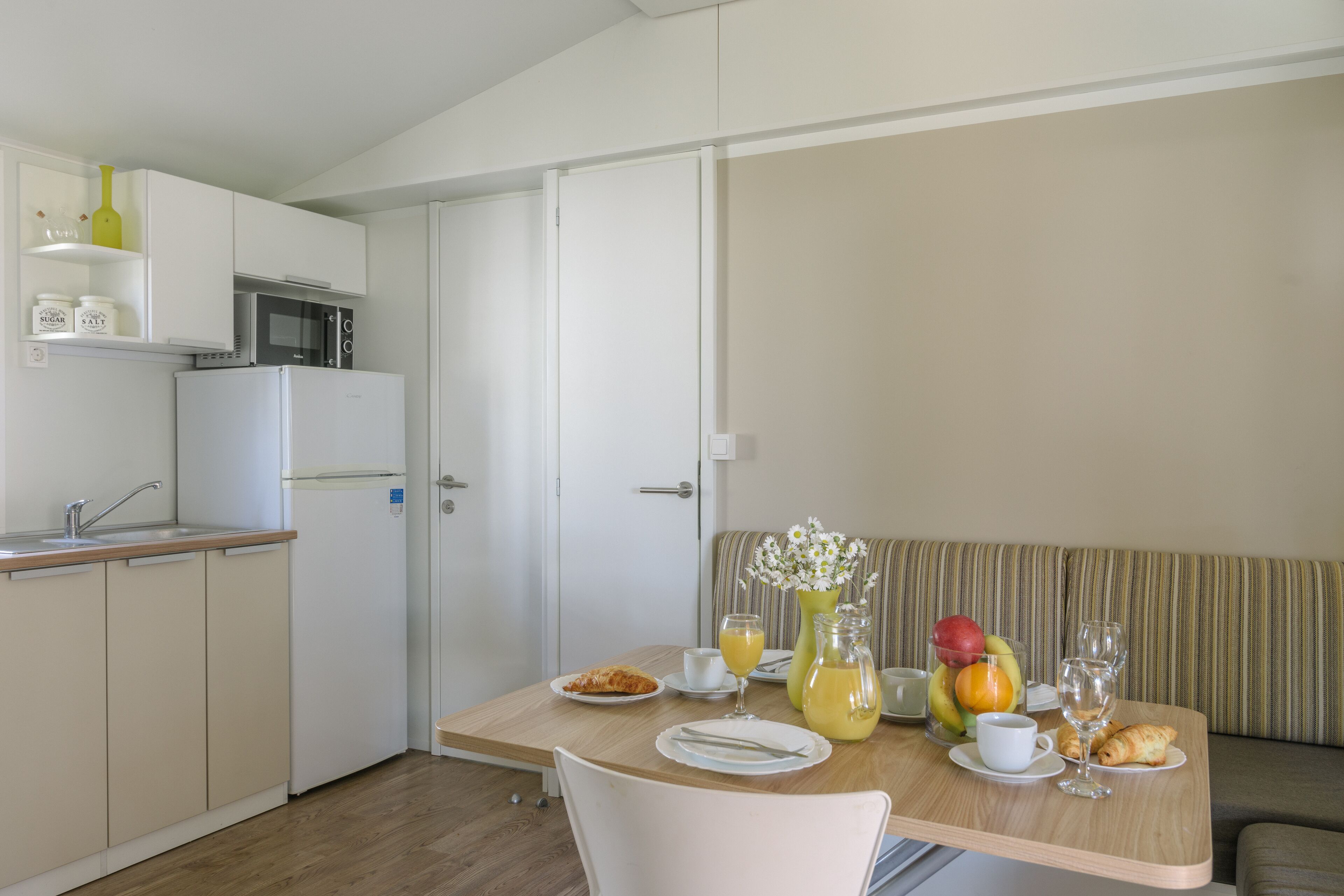 Een kitchenette met een magnetron, koelkast en een plank met potten. Een eettafel met borden, glazen en een bloevas staat voor een gestreept bankstel.