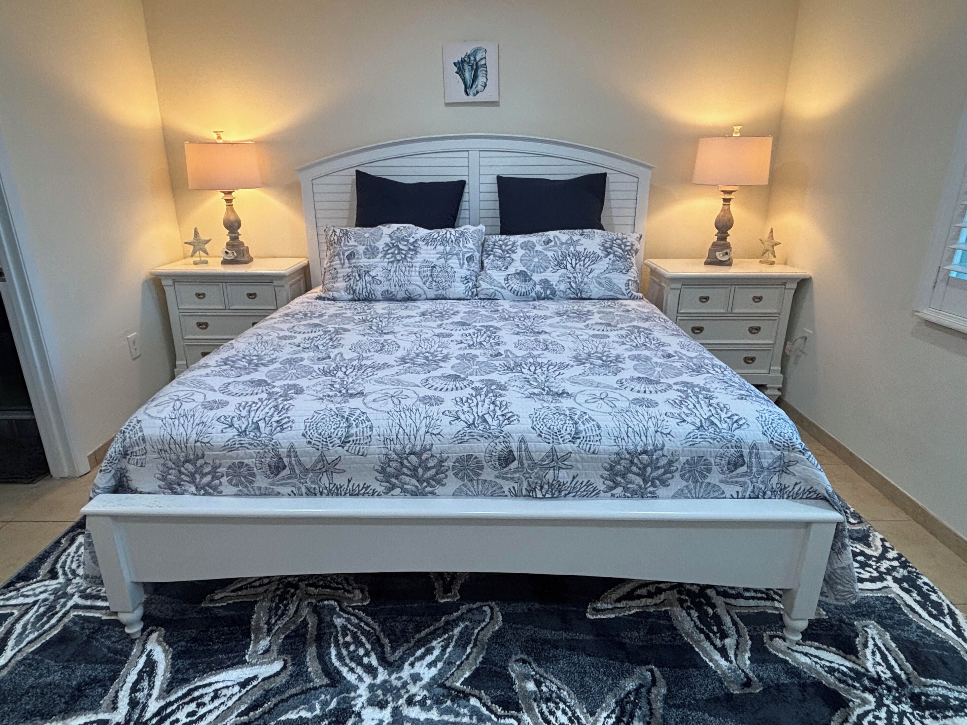 Un lit bien fait, recouvert d’une couvre-lit à motifs floraux, flanqué de deux tables de chevet avec des lampes, dans une pièce au design simple et épuré.
