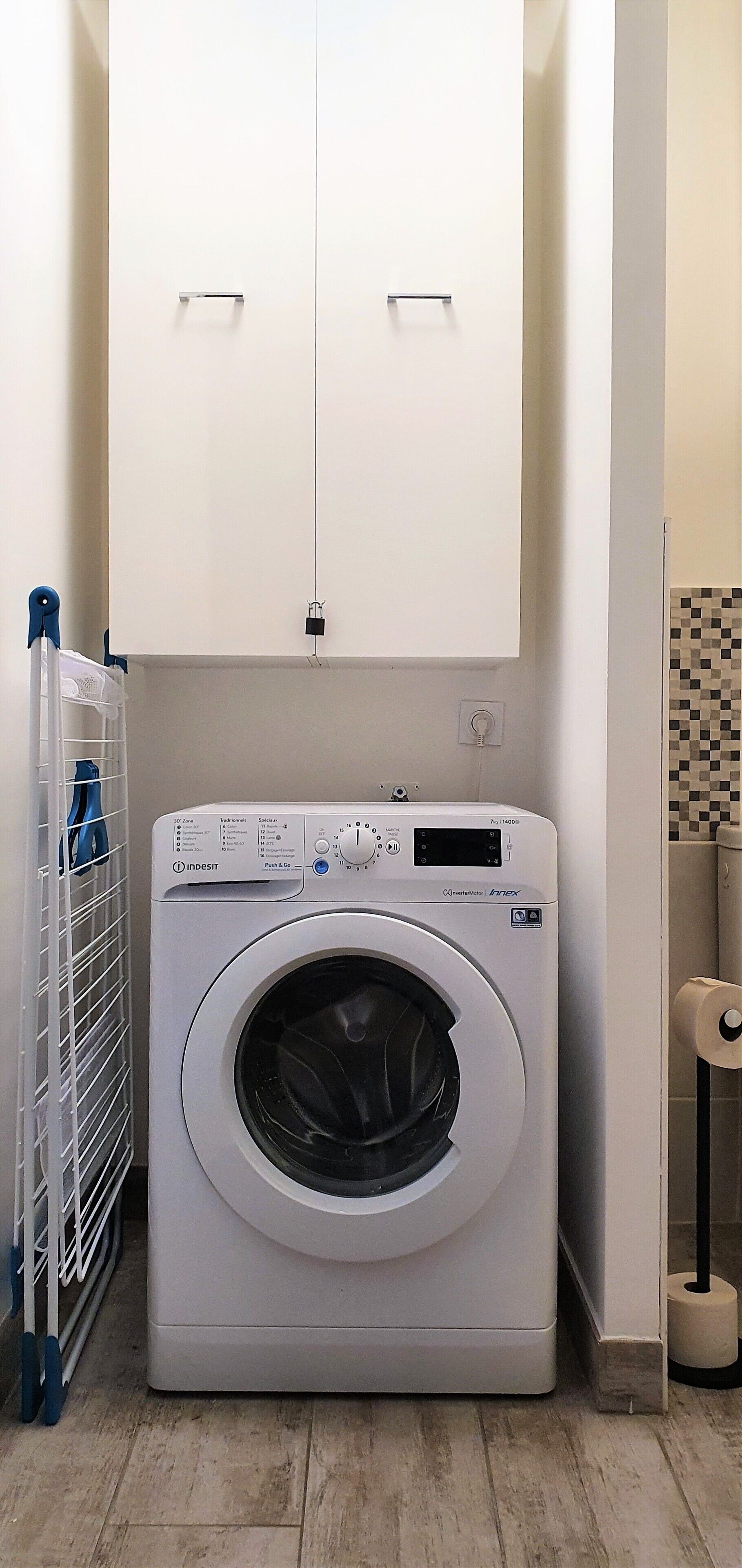 A front-loading washing machine in a laundry room with a metal drying rack and a tiled wall.