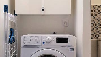 A front-loading washing machine in a laundry room with a metal drying rack and a tiled wall.