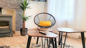 Een gezellige woonkamer met een houtkachel, een zwarte rieten fauteuil met een geel kussen, een klein tafeltje en een potplant.