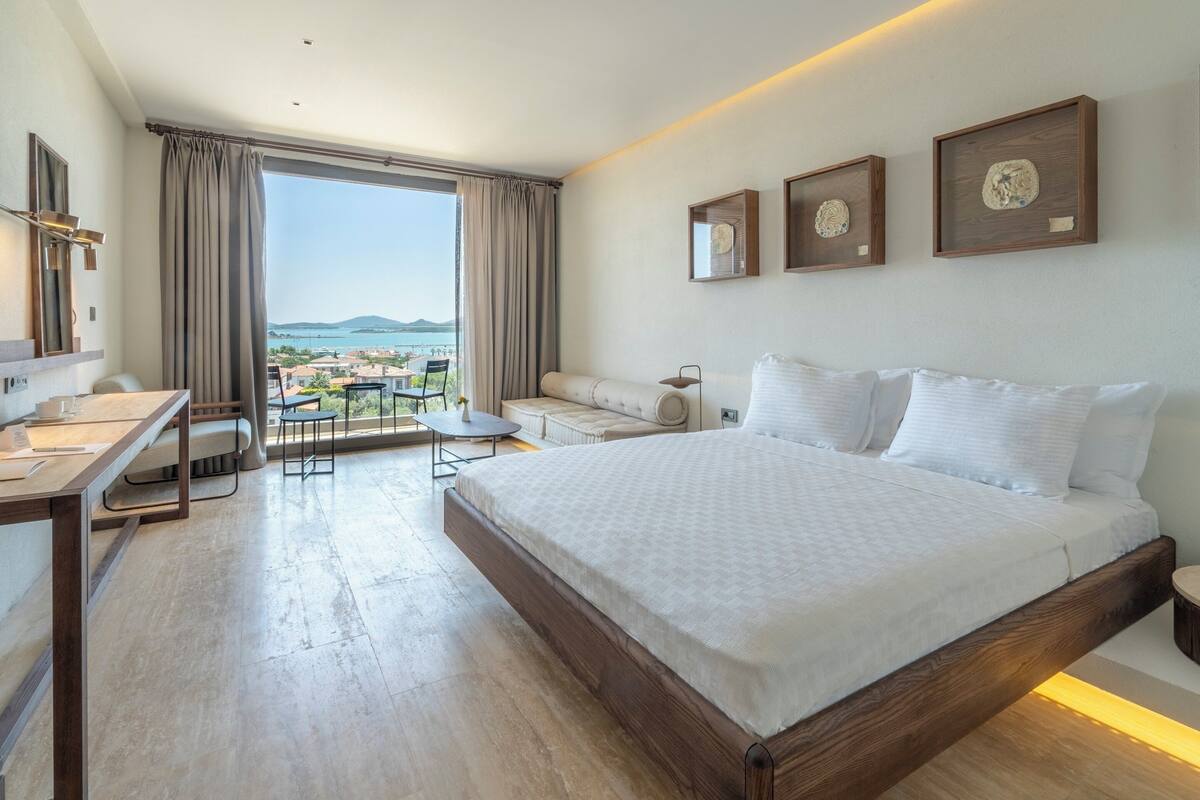 A modern hotel room with a large bed, a desk, and a balcony with a view of the sea.