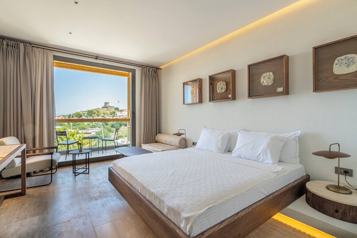 A modern hotel room with a large bed, a desk, and a balcony with a view of the city.