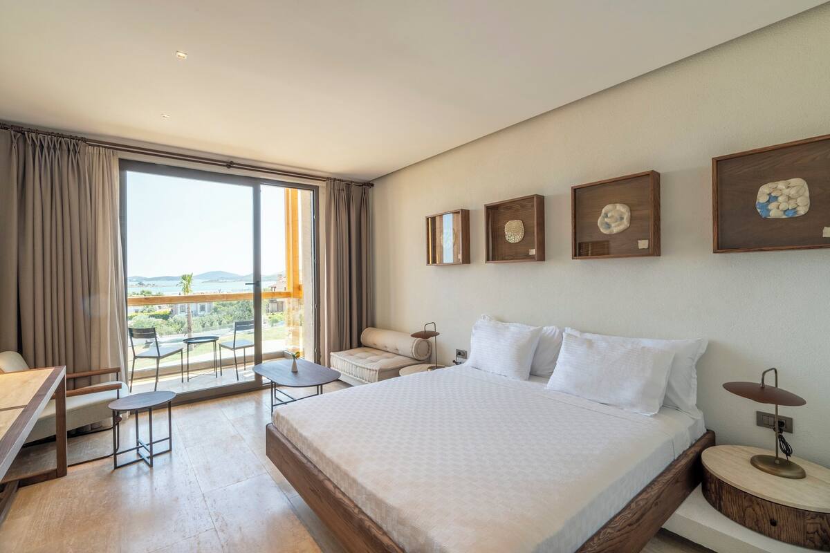 A hotel room with a large bed, a desk, a chair, a small table, and a balcony with a view.