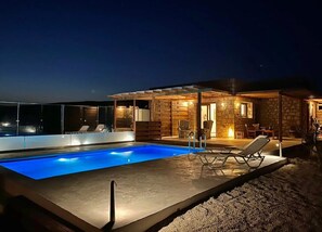 Ein moderner Poolbereich mit klarem, blauem Pool, Liegestühlen und einem Steinhaus mit überdachter Terrasse.
