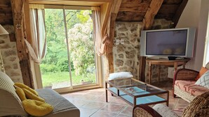 A rustic living room with a stone wall, wooden ceiling beams, a large window with curtains, a television, a sofa with yellow cushions, a wicker chair, and a small table. - Le Puy D'aubrac, Maison Familiale en Pleine Campagne (Florentin-la-Capelle)