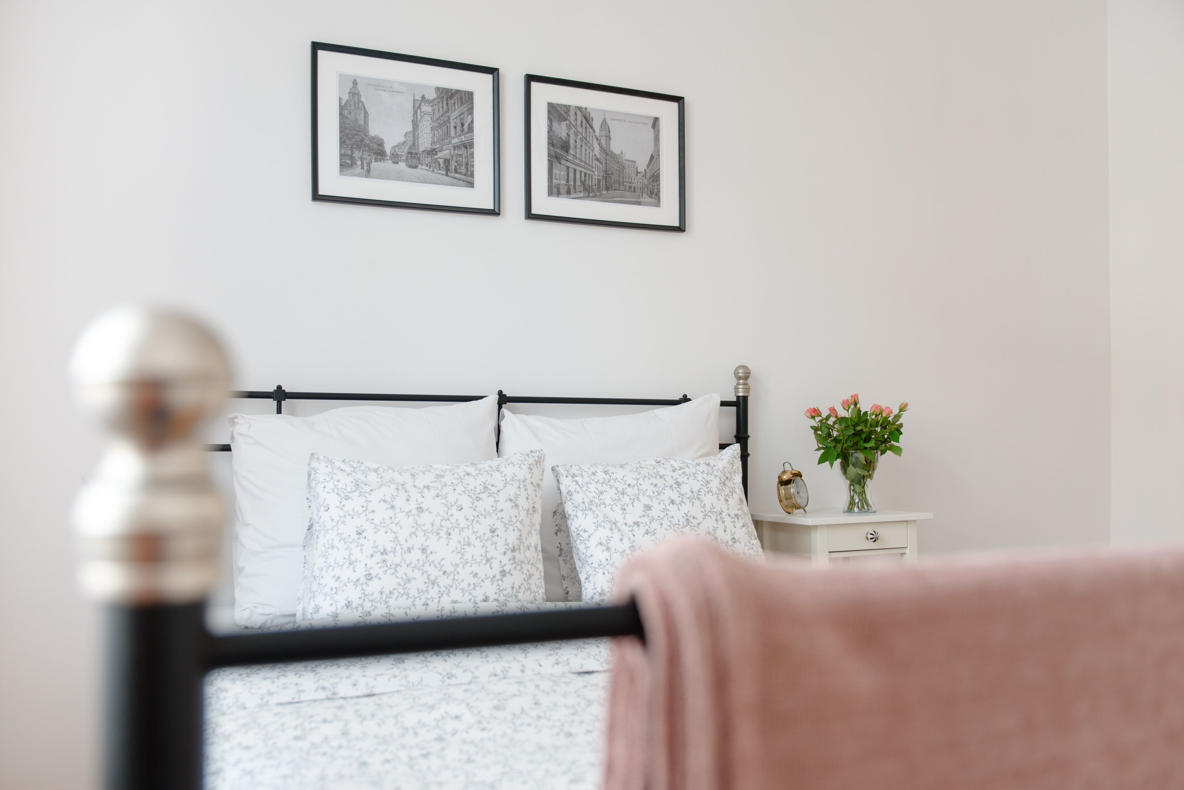 Un dormitorio con una cama, una mesita de noche con un jarrón de flores y dos cuadros en la pared.