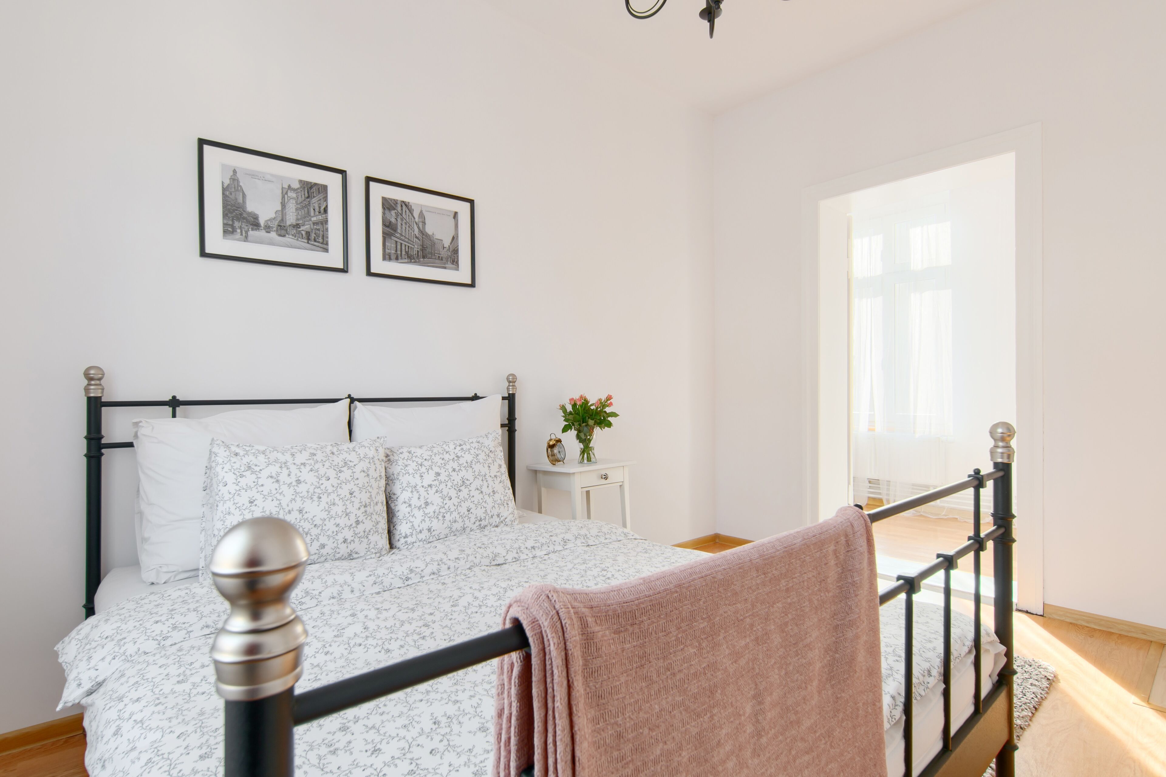Un lit bien fait, avec une structure en métal noir et du linge de lit blanc, une petite table de chevet ornée d’un vase de fleurs et deux tableaux encadrés accrochés au mur.