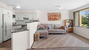 A modern kitchen with white cabinets, a dining area with a sofa and rug, and a large window with a view. - Ted's at Broulee (Broulee)