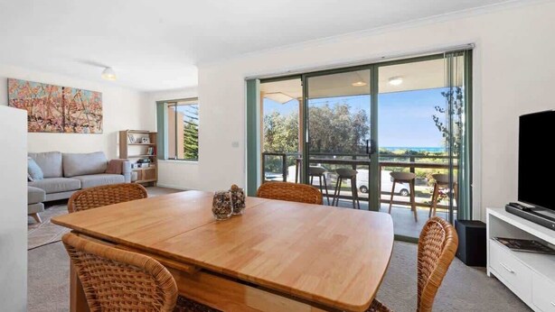 A modern living room with a wooden dining table, wicker chairs, a sofa, a TV, and a balcony with a view. - Ted's at Broulee (Broulee)