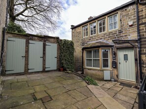 A stone courtyard with two green doors, a brick building with a bay window, and a small garden area. - Erin Cottage (Keighley)