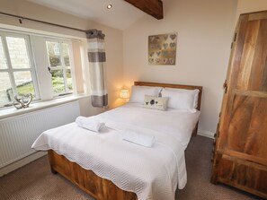 A bedroom with a wooden bed, white bedding, a window with curtains, a wall-mounted lamp, and a wooden wardrobe. - Erin Cottage (Keighley)