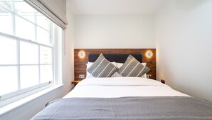 A neatly made bed with a grey blanket, a wooden headboard with two wall-mounted lights, and a window with a white blind. - 3 Bedroom flat with Patio in Central London (London)