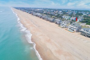 An aerial view of a beachside residential area with houses along the shoreline, featuring a clear view of the ocean and a sandy beach.