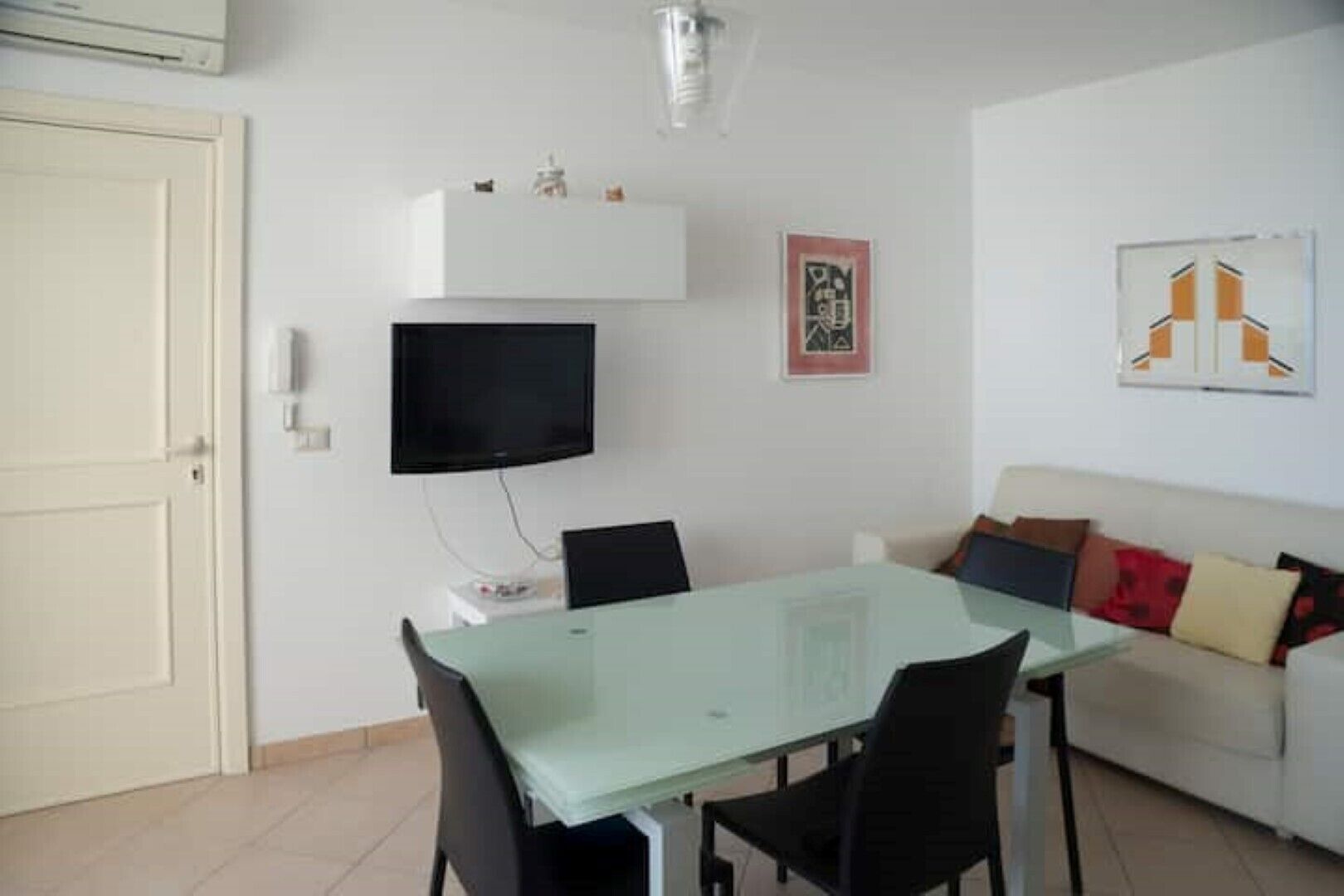 A dining area with a glass table and chairs, a flat-screen TV mounted on the wall, a white sofa with cushions, and a framed picture on the wall.