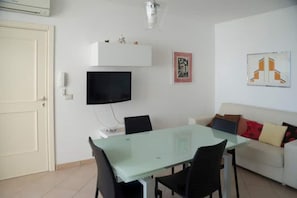Un comedor con una mesa de vidrio y sillas, un televisor de pantalla plana en la pared, un sofá blanco con cojines y un cuadro enmarcado.
