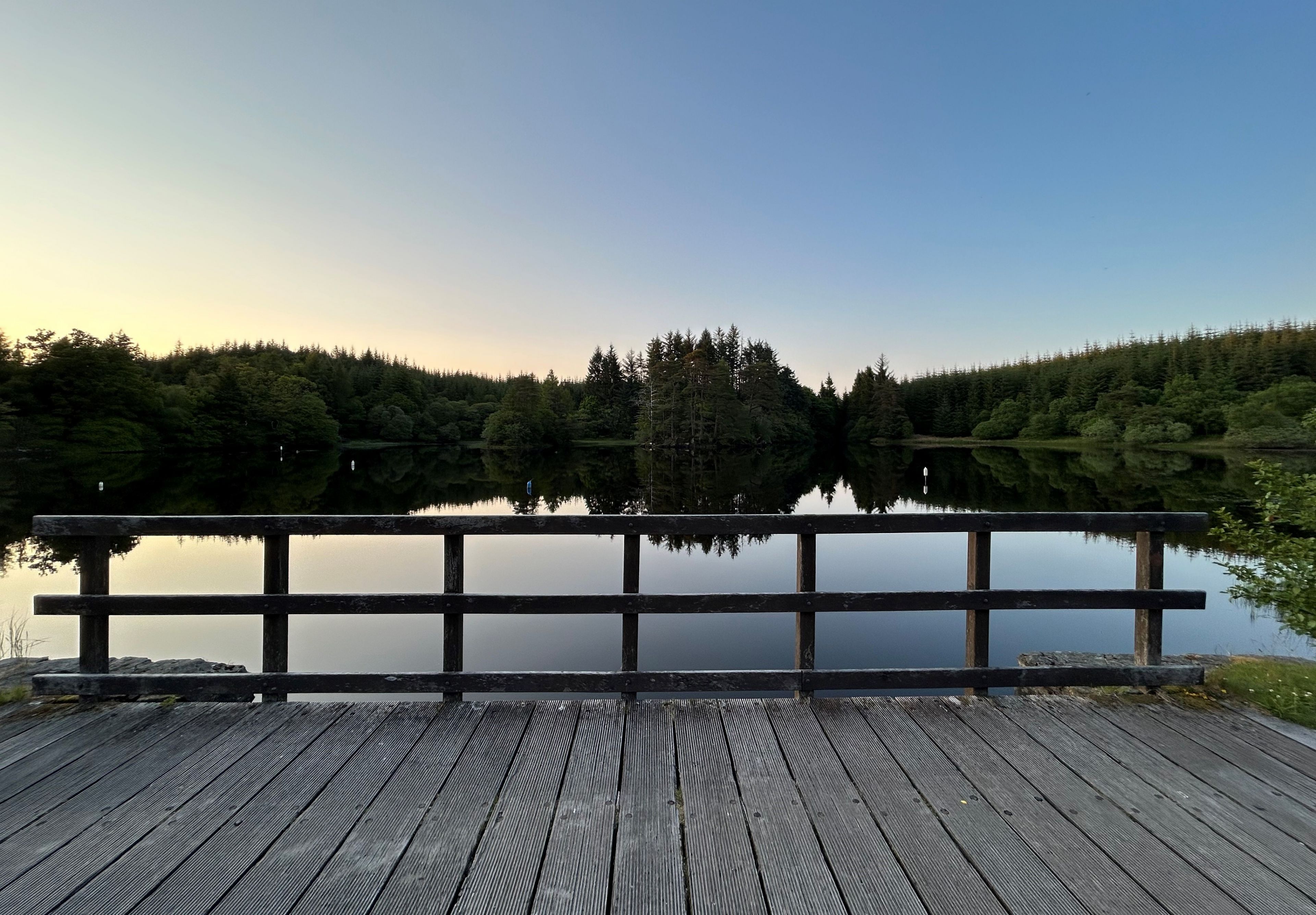 A wooden dock leading to a calm lake with a forested hill in the background.