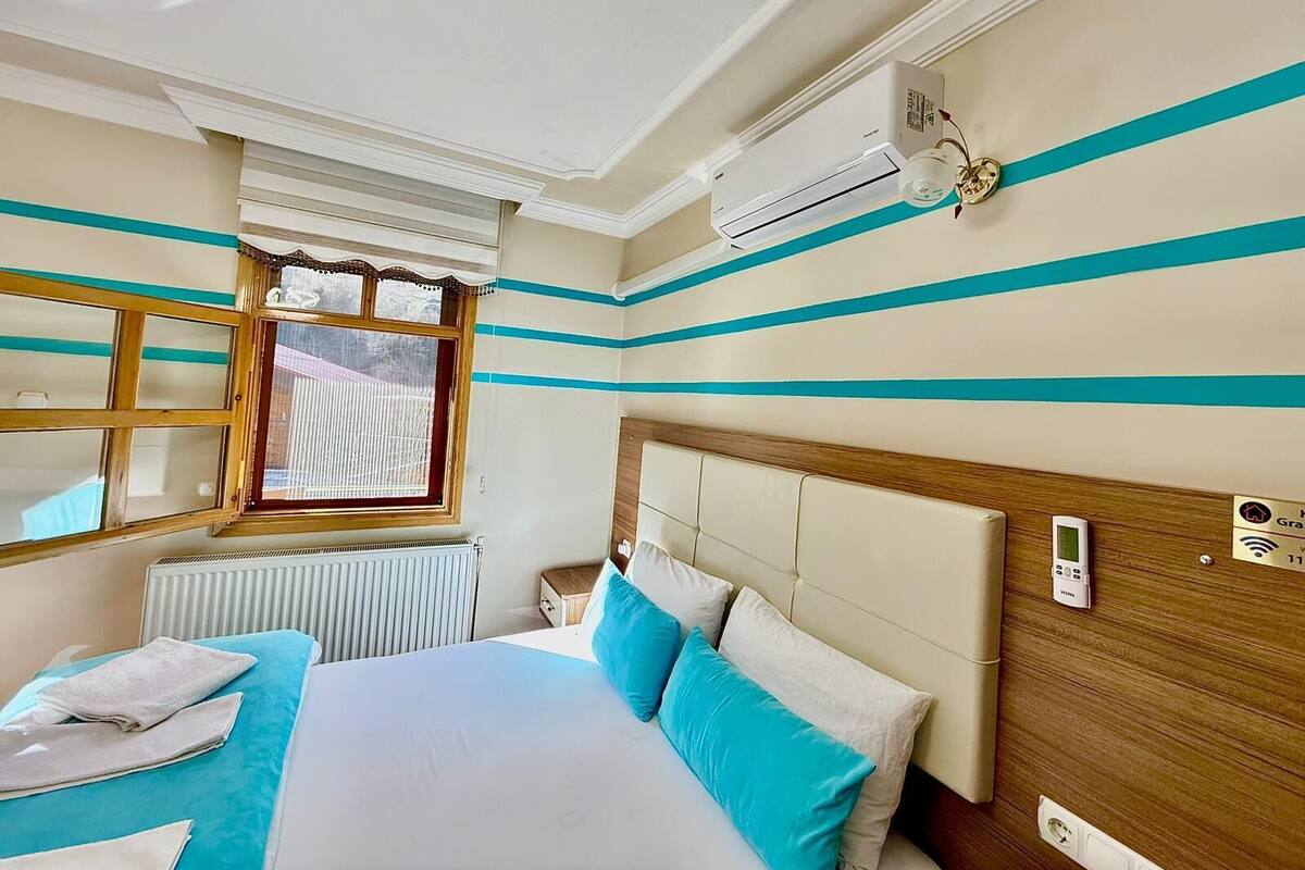 Una habitació d'hotel amb un llit, una finestra amb persianes, un aire condicionat, una taula de nit i un panell de control fixat a la paret.