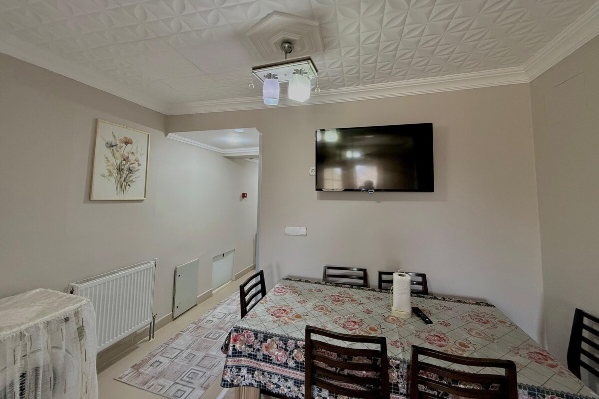 Un comedor con una mesa, sillas, un techo con diseño geométrico, un televisor montado en la pared y un radiador.