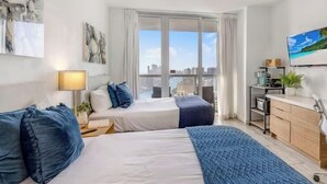 Une chambre d’hôtel équipée d’un lit, d’une table de chevet, d’une lampe et offrant une vue sur la ville grâce à de grandes fenêtres.