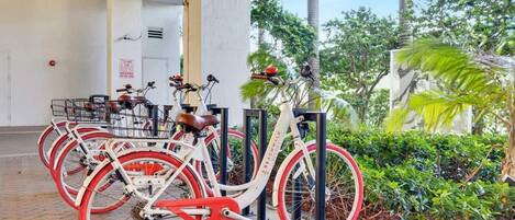 Diverses bicicletes amb pneumàtics vermells aparcades sota una zona coberta, amb palmeres i vegetació al fons.