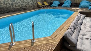 A swimming pool with a wooden deck, lounge chairs, and a cushioned seating area. - T4 Bord Canal du Midi, Piscine (Homps)