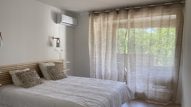 A bedroom with a bed, a bedside table, a lamp, an air conditioning unit, and a window with sheer curtains. - T4 Bord Canal du Midi, Piscine (Homps)
