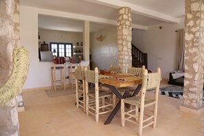 Zona pranzo con tavolo in legno e sedie intrecciate, colonne in pietra e una scala che conduce in un'altra stanza.