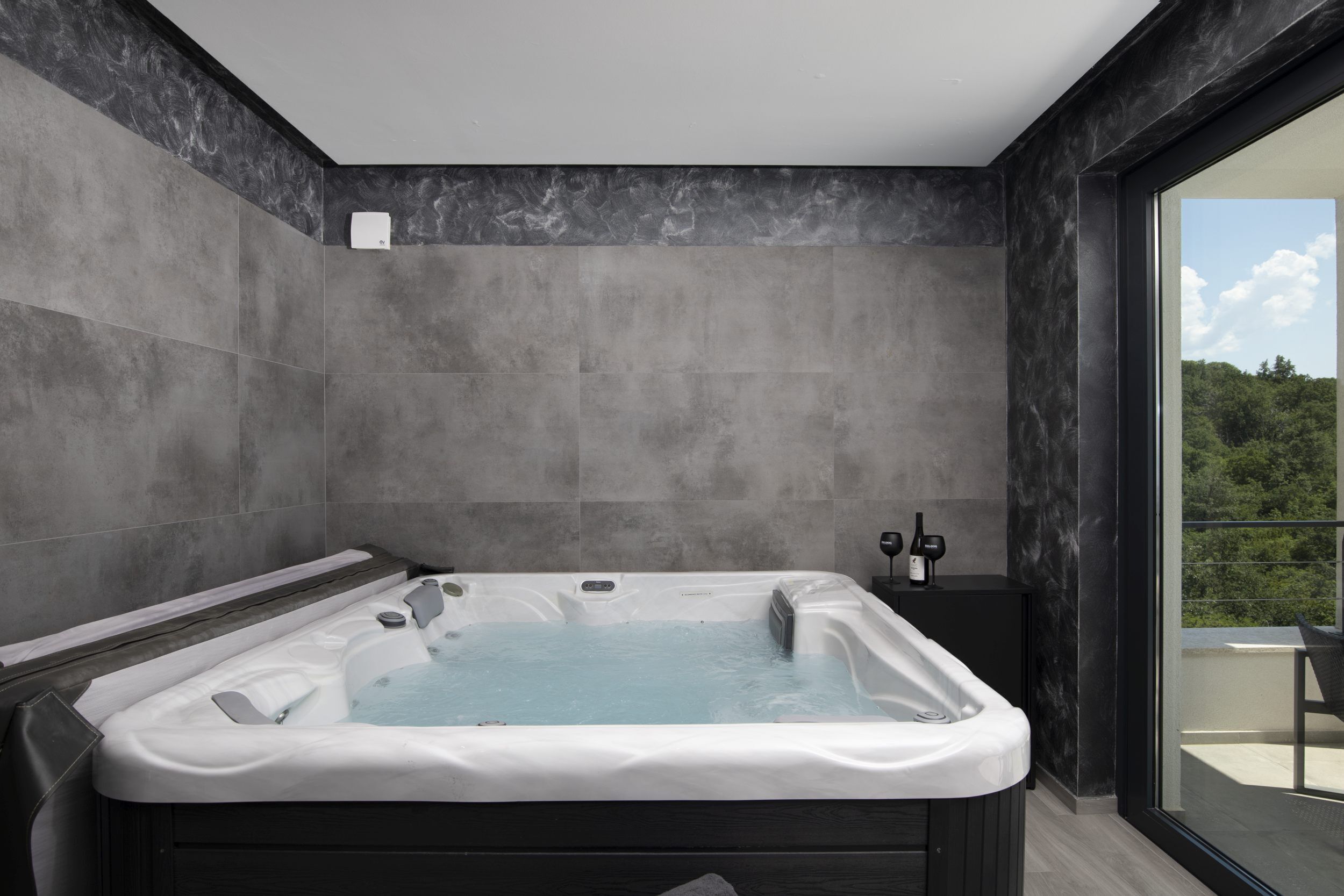 A modern bathroom with a large bathtub, a small table with a bottle, and a view of greenery through a large window.